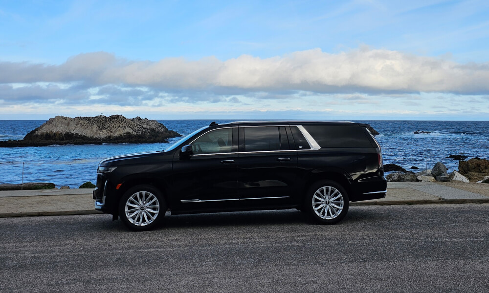 A black luxury SUV parked on a coastal road with the ocean and rocky outcrops in the background under a blue sky with streaks of white clouds